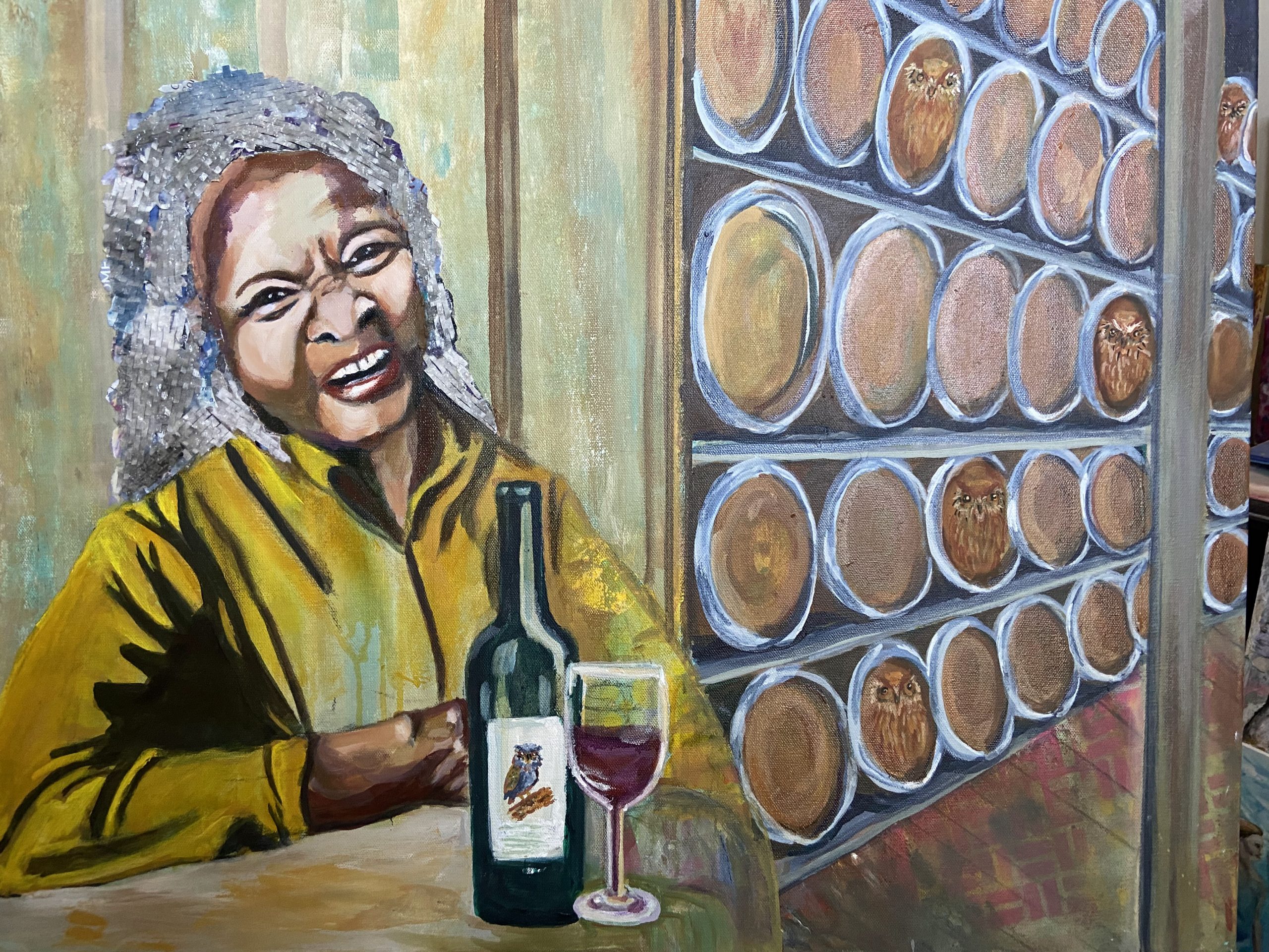 A painting titled "Barrels of Fun showing a woman sitting in a winery with a bottle of wine and a glass in front of her. 